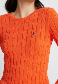 Kobieta nosząca jasnopomarańczowy sweter w warkocze z małym niebieskim haftowanym logo na lewej piersi.