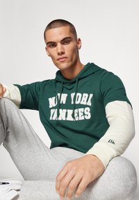 Zelená mikina s kapucňou s nápisom "NEW YORK YANKEES" v bielom písmene, v kombinácii so sivými teplákmi. Krémové akcentové rukávy; voľný strih.