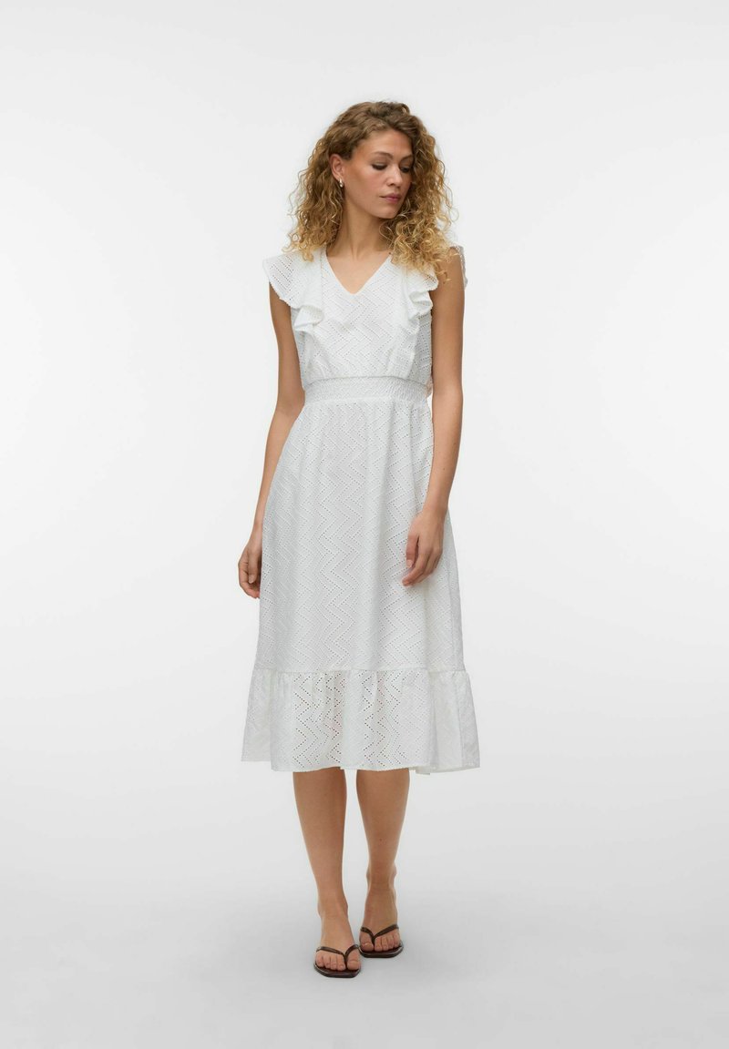Femme aux cheveux bouclés portant une robe midi blanche sans manches avec des volants et un tissu à œillets, debout pieds nus sur un fond clair.