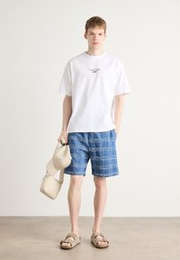 Camiseta blanca de algodón con el logo de "Calvin Klein Jeans", pantalones cortos de cuadros azules, bolso y gorra de color beige claro, sandalias marrones.