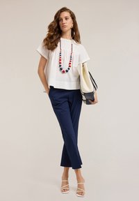 Blusa blanca de corte holgado con yugo texturizado, pantalones ajustados azul marino y sandalias blancas de tiras, complementados con un collar colorido de cuentas.