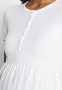 Hvid kortærmet top med en rund halsudskæring, knappelukning foran med gennemsigtige knapper og en rynket, struktureret nederste del.