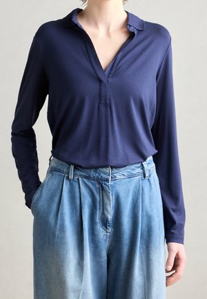 Vrouw draagt een marineblauw shirt met lange mouwen en V-hals, ingestopt in lichtblauwe hoge taille wijde jeans met handen in de zakken.