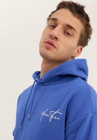 Mladý muž s krátkými hnědými vlasy, oblečený v modré mikině s bílým nápisem, dívá se mírně vzhůru na jednobarevném světlém pozadí.