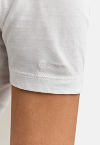 Gros plan sur le bras d'une personne portant un t-shirt blanc à manches courtes avec un logo "GANT" subtilement embossé près de l'ourlet.