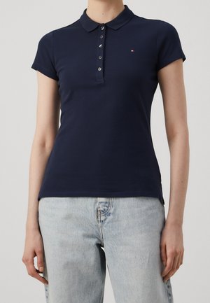 Femme portant un polo bleu marine à manches courtes avec des boutons et un jean bleu clair sur un fond uni.