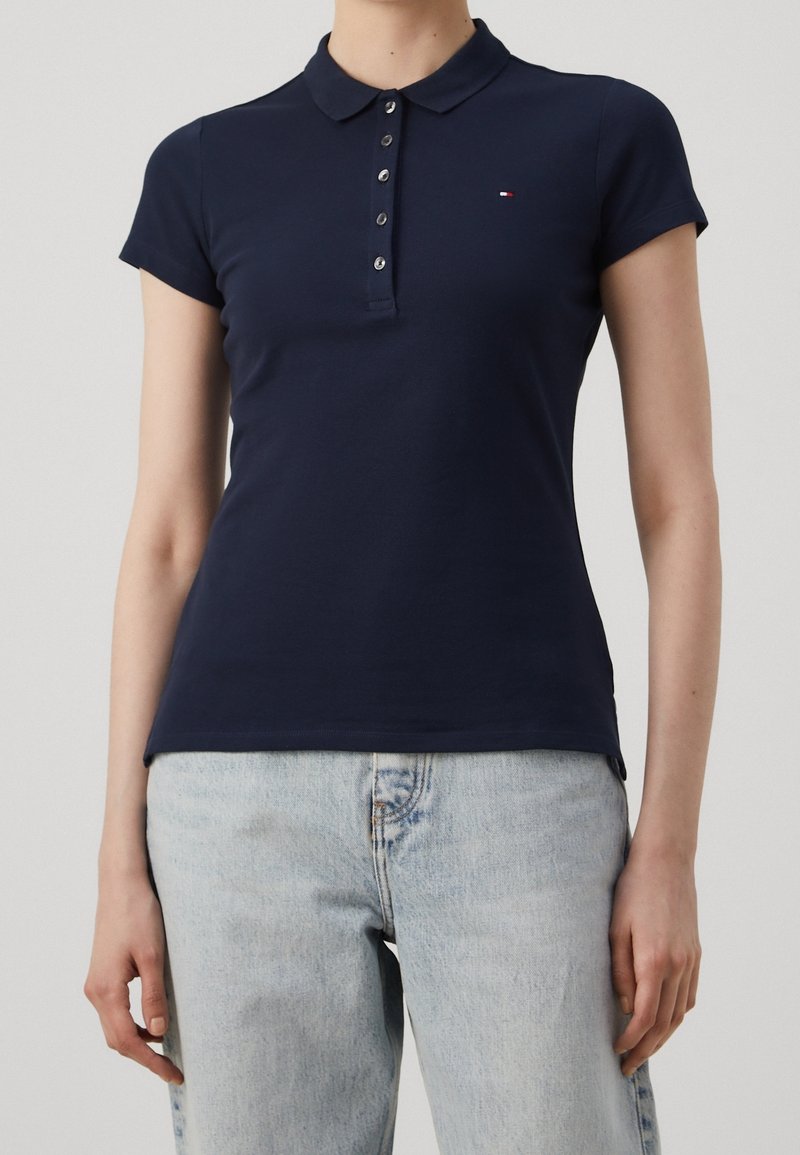 Femme portant un polo bleu marine à manches courtes avec des boutons et un jean bleu clair sur un fond uni.