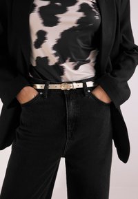 Person trägt schwarze Jeans mit weißem Gürtel, schwarz-weiß gemustertes Top und schwarze Jacke, Hände in den Taschen.