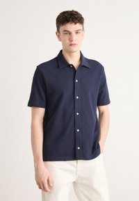 Camicetta a maniche corte blu navy con colletto appuntito, caratterizzata da una chiusura frontale con bottoni e un tessuto leggermente texturizzato. Bottoni bianchi che migliorano il contrasto.