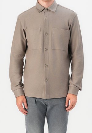 Mężczyzna ubrany w taupe koszulę na guziki z długim rękawem i dwoma kieszeniami na piersi oraz szare spodnie, stojący na jednolitym tle.