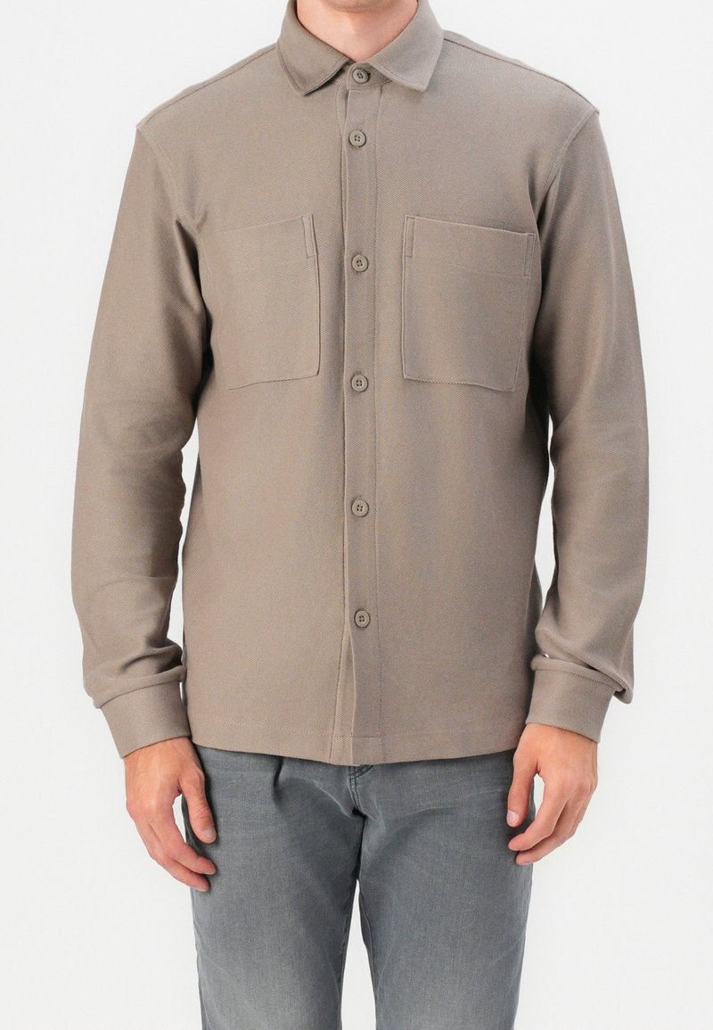 Homme portant une chemise taupe à manches longues avec boutons et deux poches sur la poitrine, ainsi qu'un pantalon gris, debout devant un fond uni.