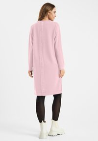Vrouw in een roze jurk met lange mouwen en verticale logotekst op de rug, zwarte panty, en witte chunky enkellaarsjes, staand met de rug naar ons gekeerd.