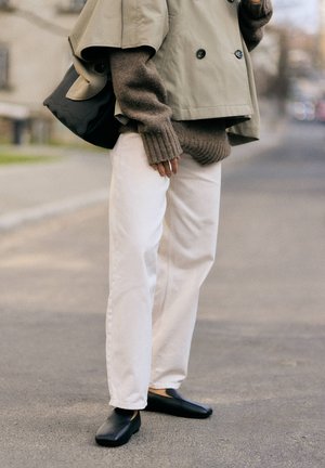 Person steht auf dem Gehweg, trägt eine beige kurze Jacke, einen braunen Strickpullover, weiße Hose, schwarze Schuhe und trägt eine schwarze Handtasche.