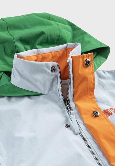 Col de veste avec capuche verte, extérieur gris clair, doublure intérieure orange, fermeture éclair partiellement ouverte et boutons-pression sur la patte orange.