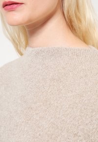 Hellbeiger Pullover aus strukturiertem Stoff mit hohem Kragen, der ein sanftes, gemütliches Aussehen und minimale Nähdetails zeigt.