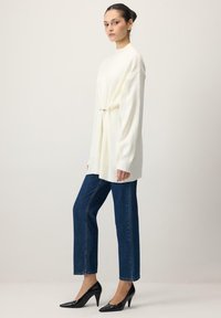 Valge kudumissärk, millel on üle mõõdukad varrukad ja mis on vööl hõbedase klambriga kitsaks tõmmatud; kombineeritud siniste sirgete teksade ja mustade teravate kontsadega.