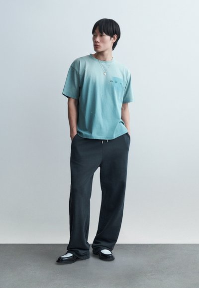 Νέος άνδρας που φοράει χαλαρό τιρκουάζ μπλουζάκι, μαύρο παντελόνι με φαρδιά μπατζάκια, μαύρα και λευκά παπούτσια, στέκεται με τα χέρια στις τσέπες μπροστά σε ένα απλό φόντο.