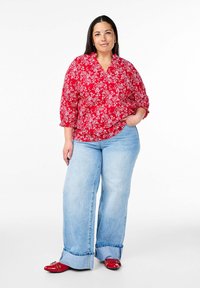 Blusa rossa a fiori con maniche 3/4, abbinata a jeans a gamba larga di colore blu chiaro e scarpe rosse con decorazioni. Texture lisce in tutto.