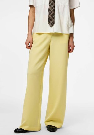 Osoba ubrana w luźne jasnopopielate spodnie, białą koszulę z krótkim rękawem, brązowy kratkowany krawat i czarne buty wsuwane, stojąca na białym tle.