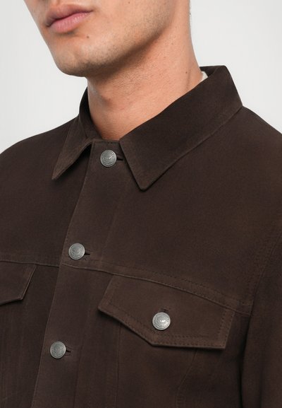 Veste marron en tissu doux, avec un col pointu, deux poches avant et des boutons-pression en métal argenté.