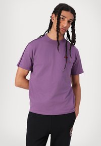Camiseta de algodón morada con mangas cortas, cuello redondo y pequeño detalle de logo rojo en el pecho. Se lleva con pantalones negros de textura suave.