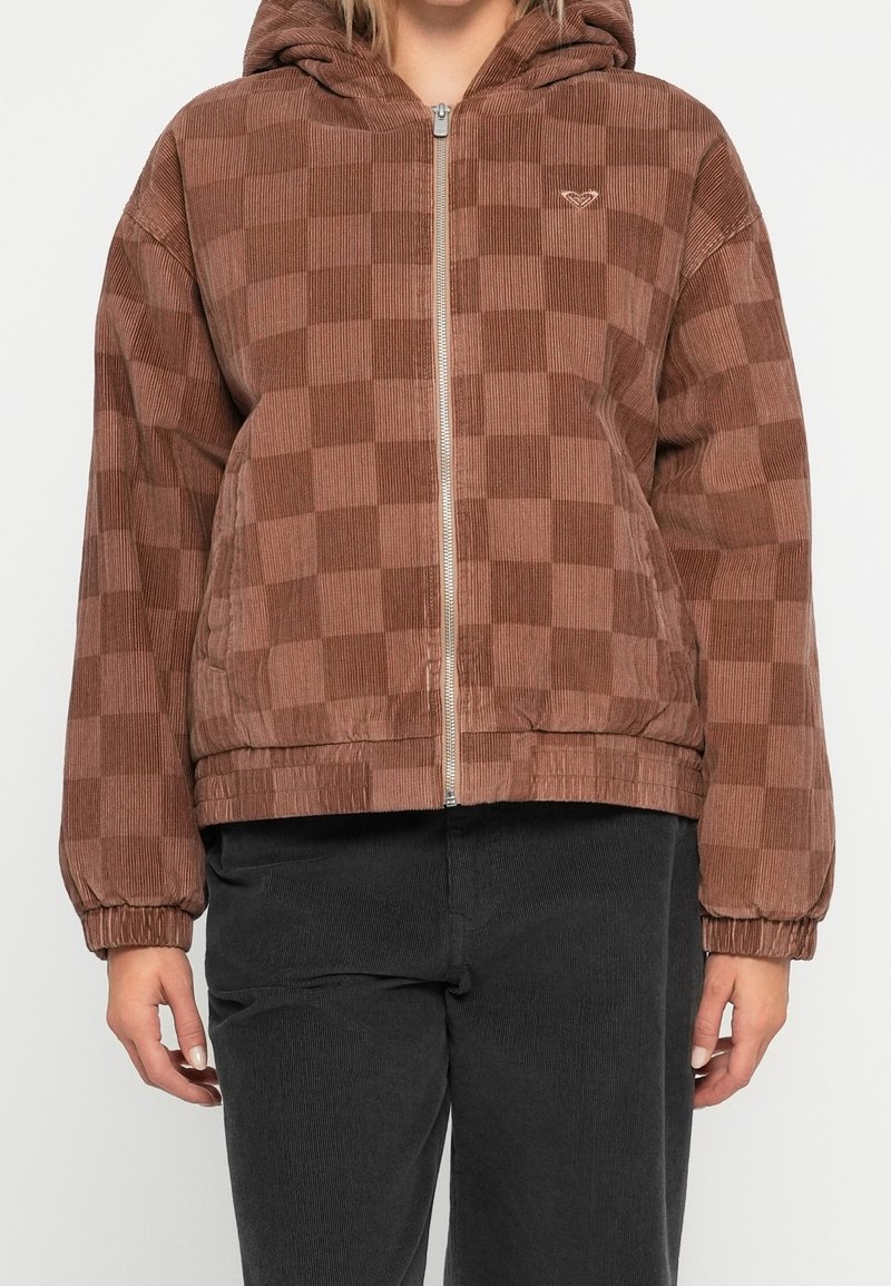 Personne portant une veste à capuche zippée marron à carreaux avec des poignets élastiques et un pantalon noir, debout devant un fond uni.