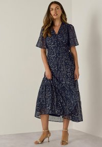 Abito blu navy con maniche corte a campana, caratterizzato da un motivo a pois bianchi, un design avvolgente nella parte anteriore e una gonna a strati. Indossato con tacchi beige.