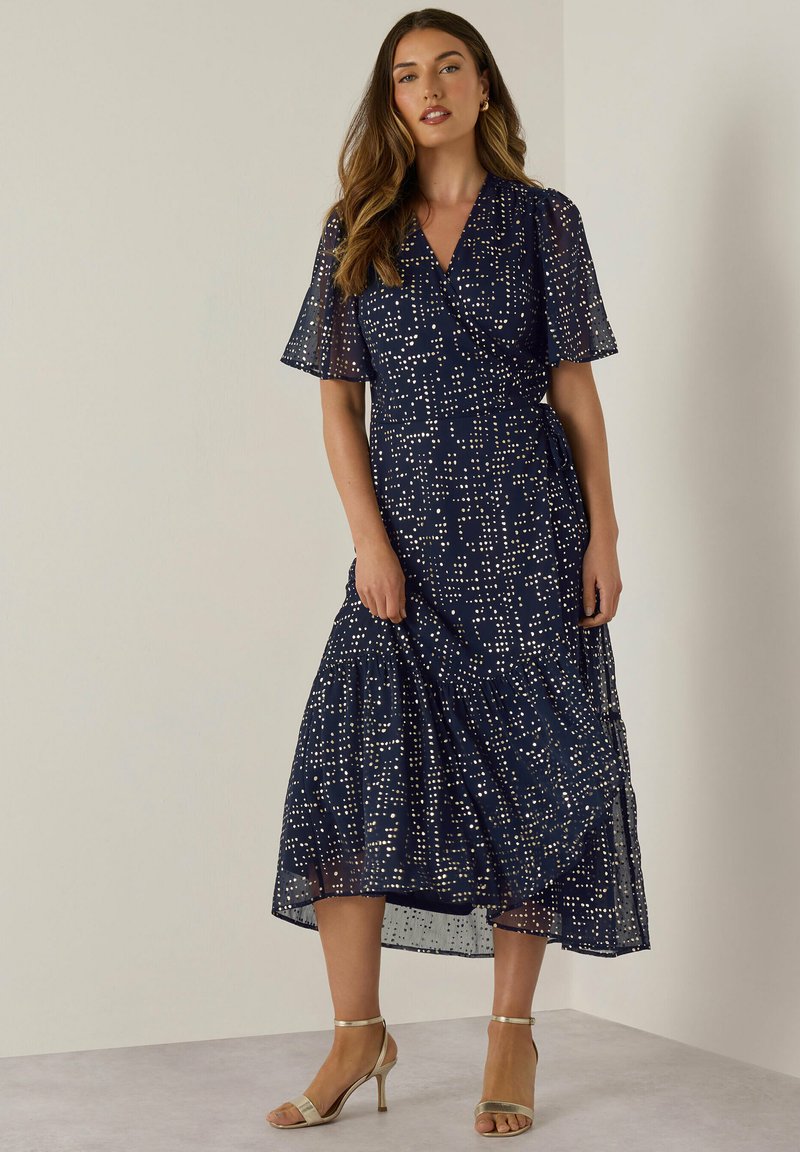 Abito blu navy con maniche corte a campana, caratterizzato da un motivo a pois bianchi, un design avvolgente nella parte anteriore e una gonna a strati. Indossato con tacchi beige.