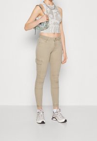 Kvinna klädd i beige cargojoggingbyxor, grå grafisk croptop, vita strumpor, vita sneakers och håller en liten silverfärgad axelväska.