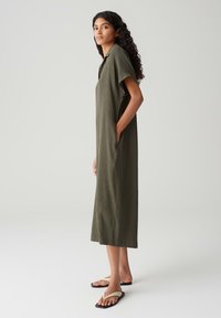 Long, olive-green dress with a relaxed fit, short sleeves, textured fabric, and side pockets. Worn with simple black sandals.