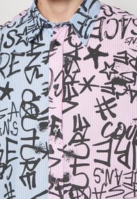 Camisa de botón con un diseño partido: la izquierda en rayas verticales azules, la derecha en rosa con texto y formas en negrita estilo grafiti en negro.