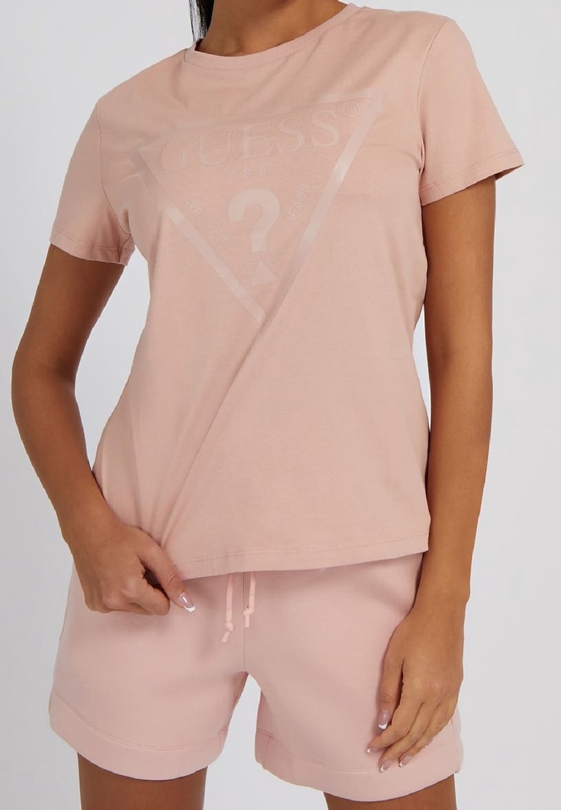 Femme portant un t-shirt Guess rose clair avec logo et un short assorti, debout, tenant l'ourlet de son t-shirt d'une main.