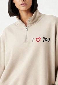 Pull-over beige avec fermeture quart de zip en tissu doux. Présente un cœur rouge et un texte noir "I LOVE YOU" brodés sur le côté gauche de la poitrine.