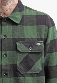 Grön och svart rutig skjorta i flanell med knäppkrage, bröstficka och svarta knappar. Har ett texturerat tygmönster.