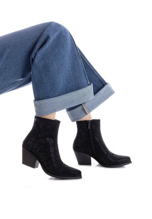 Botas de tobillo negras con tacón, con una superficie texturizada y un patrón geométrico, con cremallera lateral, combinadas con unos jeans de mezclilla azul con dobladillo.