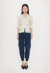 Beige cropped shirt with puff sleeves, featuring two chest pockets and a front zipper, paired with blue denim jogger pants, and black pointed shoes.