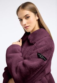 Fioletowy futrzany płaszcz z szerokim kołnierzem i długimi rękawami. Posiada metkę z logo na rękawie. Miękka tekstura i objętościony design.