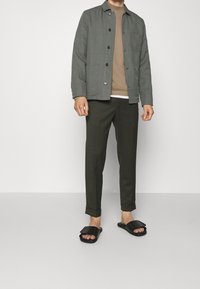 Uomo con giacca grigia chiusa da bottoni, maglione beige, pantaloni verde scuro con risvolto e sandali neri, in piedi su uno sfondo neutro.
