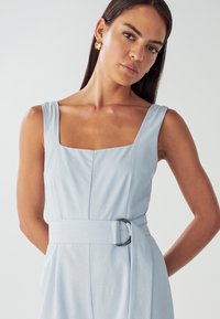 Robe bleu clair avec un décolleté carré, des bretelles larges et une ceinture avec une boucle circulaire en argent. Tissu lisse, coupe ajustée.