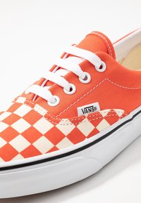Orange og kremfarvede ternede canvas Vans sneakers med hvide snørebånd, sort sål og logo-mærke; har en flad design og tekstureret overdel.