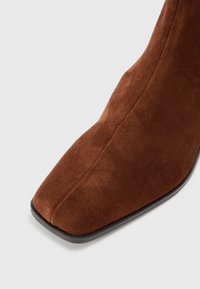 Bocanci scurti din suede maro cu vârf pătrat și cusături vizibile pe un fundal deschis.