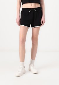 Pantaloni corti sportivi neri con vita regolabile, caratterizzati da una rifinitura bianca lungo l'orlo. Indossati con scarpe da ginnastica bianche e calze chiare.