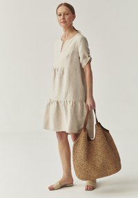 Robe en lin beige clair avec un col rond, des manches courtes et un détail de taille froncée. Tient un grand sac en paille tressée avec des accents en cuir.