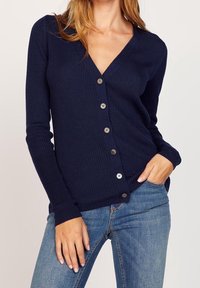 Kvinna klädd i en marinblå ribbad kofta med knappar och ljusblå jeans, ena handen i fickan och den andra vilande på benet.