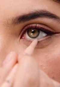 Gros plan sur un œil avec une iris marron clair, un eyeliner défini, et un bout de doigt appliquant un crayon près de la ligne des cils inférieure. La peau semble lisse.