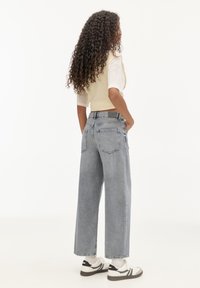 Jean denim gris taille haute à jambes larges avec deux poches arrière et ourlets effilochés ; stylisé avec un gilet beige court et une chemise blanche.