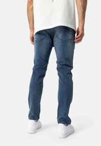 Jeans de mezclilla azul oscuro con un corte recto, que cuentan con bolsillos traseros y una textura de desvanecimiento sutil. Combinados con zapatillas blancas.