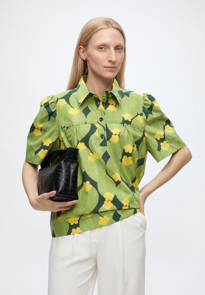 Femme aux cheveux blonds et raides, portant un chemisier vert à motifs floraux et un pantalon blanc, tenant un sac à main noir texturé sur un fond uni.