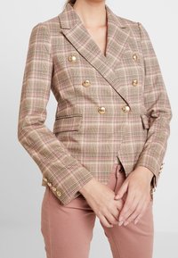 Donna che indossa un blazer doppiopetto a quadri beige con bottoni dorati e pantaloni rosa polveroso, mani unite davanti.