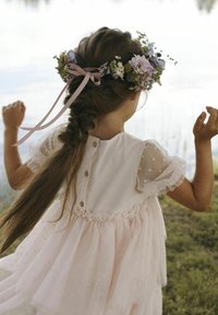 Robe rose pâle avec un ourlet volanté et des manches courtes en voile, décorée de petits pois ; une couronne de fleurs avec des rubans orne les cheveux.
